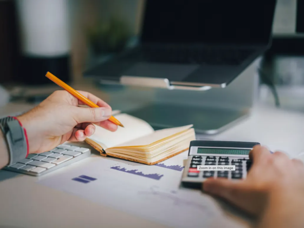 Hands using a calculator and pencil beside an open notebook and printed charts.