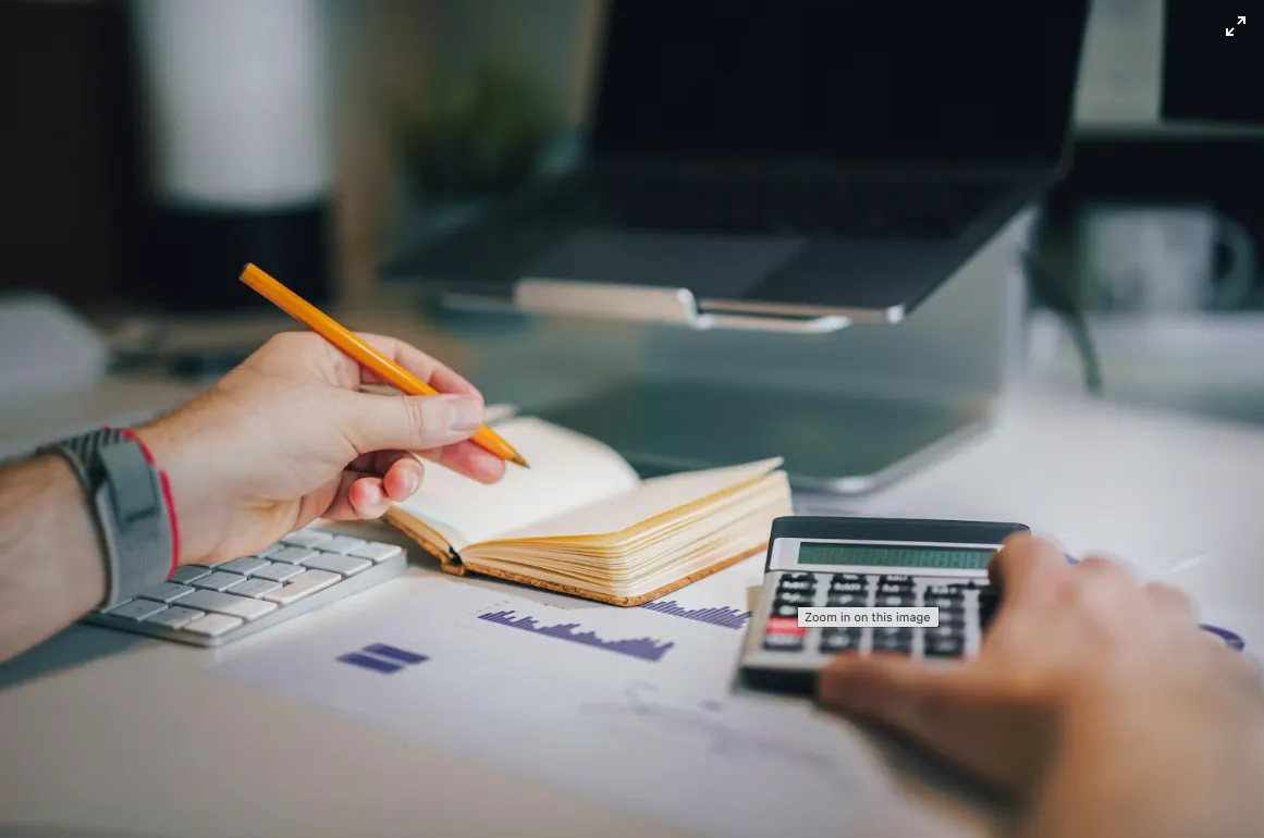 Hands using a calculator and pencil beside an open notebook and printed charts.